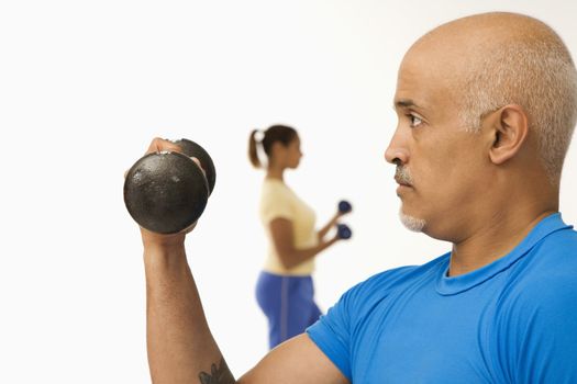 Close up of mid adult multiethnic man exercising using dumbbells with mid adult multiethnic woman in background.