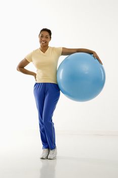 Mid adult multiethnic woman standing and holding blue exercise ball looking at viewer and smiling.