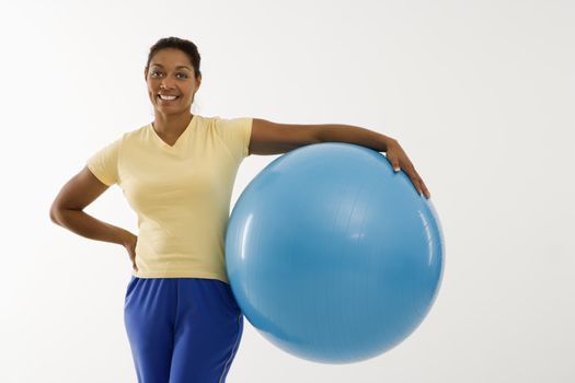 Mid adult multiethnic woman standing and holding blue exercise ball looking at viewer and smiling.