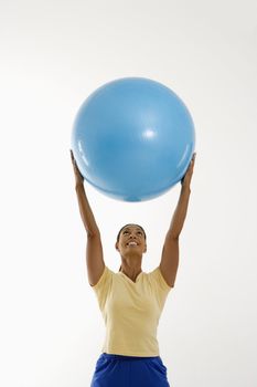 Mid adult multiethnic woman standing and holding blue exercise ball over her head smiling.