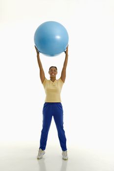 Mid adult multiethnic woman standing and holding blue exercise ball over her head looking at viewer and smiling.