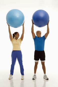 Mid adult multiethnic man and woman holding blue exercise balls over their heads looking at each other smiling.