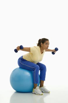 Mid adult multiethnic woman balancing on blue exercise ball with outstretched arms holding dumbbells.