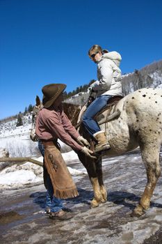 Caucasian male wrangler helping young adult Caucasian female on horseback.