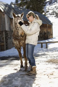 Young adult Caucasian woman petting horse with stable in background in winter.