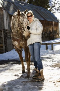 Young adult Caucasian woman petting horse with stable in background in winter.