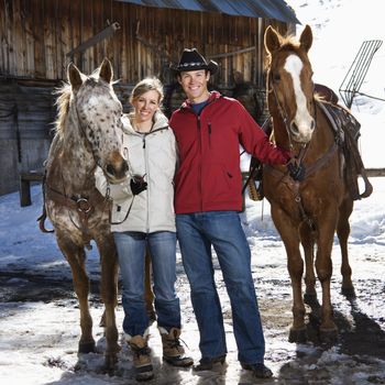 Caucasian couple holding horses in winter with stable in background.