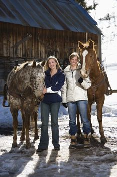 Two Caucasian women holding horses smiling at viewer with stable in background.