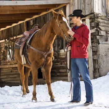 Young Caucasian man in western attire petting his horse with stable in background.