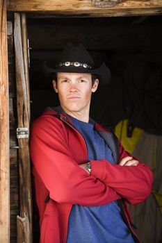 Young adult Caucasian male wearing cowboy hat with arms crossed leaning and looking at viewer.