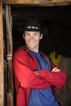 Young Caucasian man wearing cowboy hat leaning in doorway with arms crossed smiling at viewer.