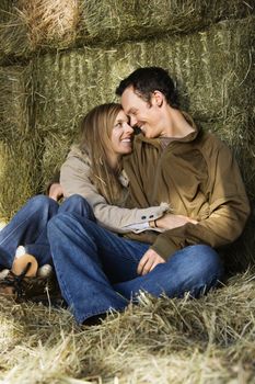 Young adult Caucasian couple sitting on hay hugging and smiling at each other.