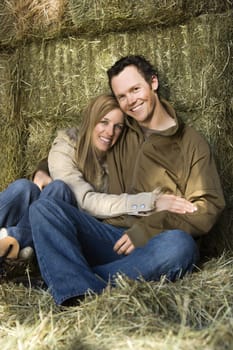 Young adult Caucasian couple sitting on hay hugging and smiling at viewer.