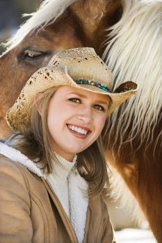Young Caucasian woman wearing cowboy hat and smiling at viewer next to horse.