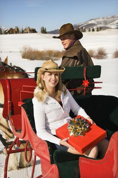 Young Caucasian woman holding a present while man drives horse-drawn sleigh.