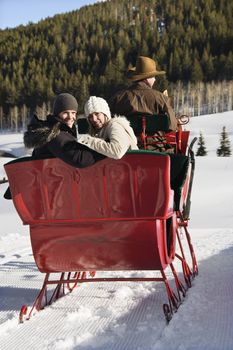 Rear view of man driving horse drawn sleigh with young couple looking back at viewer.