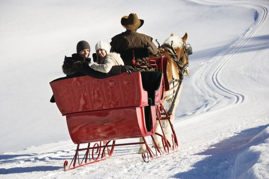 Rear view of man driving horse drawn sleigh with young couple looking back at viewer.