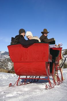 Rear view of young Caucasian couple and mid-adult man looking over shoulder at viewer from horse drawn sleigh.