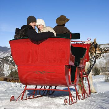 Rear view of man driving horse drawn sleigh with young couple looking back at viewer.