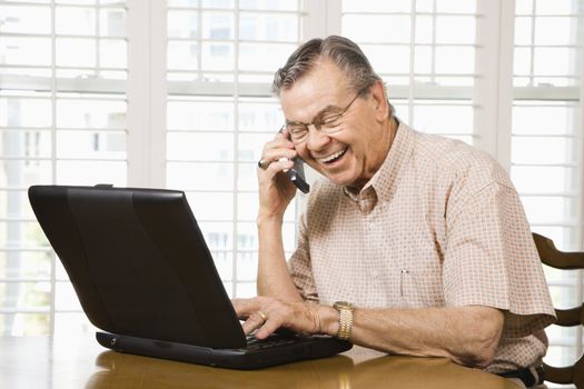 Mature Caucasian man typing on laptop and talking on cellphone.