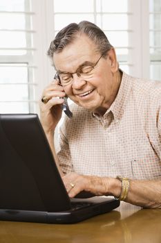 Mature Caucasian man typing on laptop and talking on cellphone.