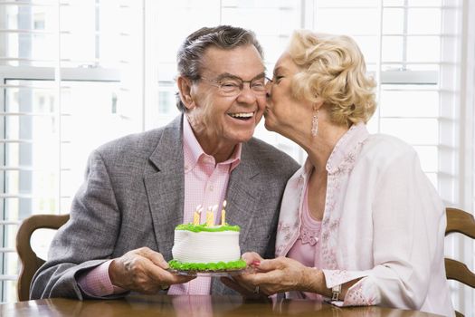 Mature Caucasian woman kissing mature Caucasian man while holding birthday cake.