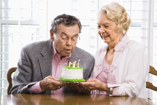 Mature Caucasian man blowing candles out on birthday cake while Mature Caucasian woman watches.