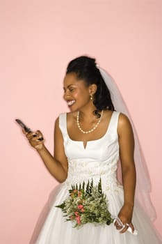 Mid-adult African-American bride looking at cellphone on pink background.