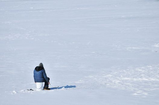 Ice fisherman sitting on a bucket above hole in the ice.