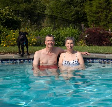 Senior couple relax in pool with pet dog on the edge of the pool