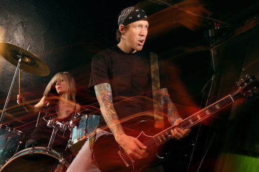 Band playing on a stage. Female drummer and male guitarist. Shot with strobes and slow shutter speed to create lighting atmosphere and blur effects. Slight motion blur on performers.