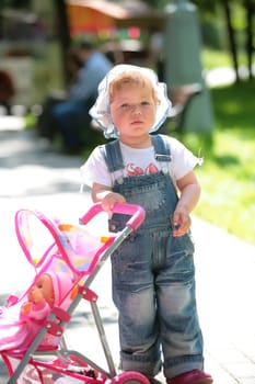 small girl in jeans overalls walks in park with doll in sidercar