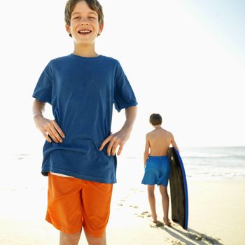 Caucasian boy with hands on hips with another Caucasian boy with bodyboard in background.