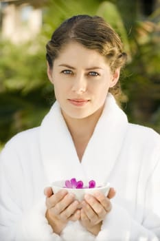 Attractive Caucasian mid-adult woman in white robe holding bowl of purple orchids floating in water.