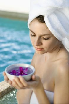 Caucasian mid-adult woman wearing white terry robe holding bowl of purple orchids next to pool.