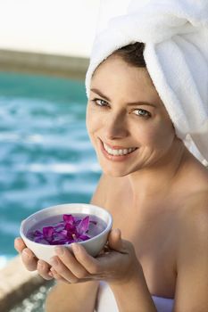 Caucasian mid-adult woman wearing towel around head and body holding bowl of purple orchids next to pool.