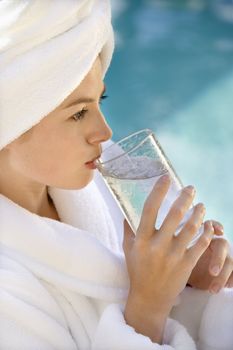 Caucasian mid-adult woman wearing white terry robe drinking water next to pool.