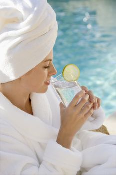 Caucasian mid-adult woman wearing robe and towel on head drinking from glass next to pool.