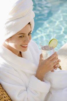 Caucasian mid-adult woman wearing robe and towel on head drinking from glass next to pool.