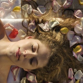 Above view of bare Caucasian mid-adult woman lying down with eyes closed and hair spread out surrounded by rose petals.