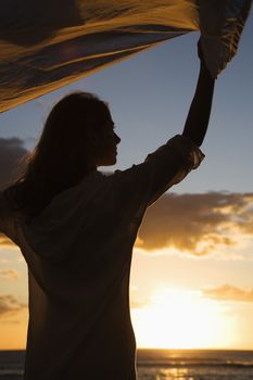 Attractive Caucasian mid-adult woman holding up fabric in breeze silhouetted by sunset beside ocean.