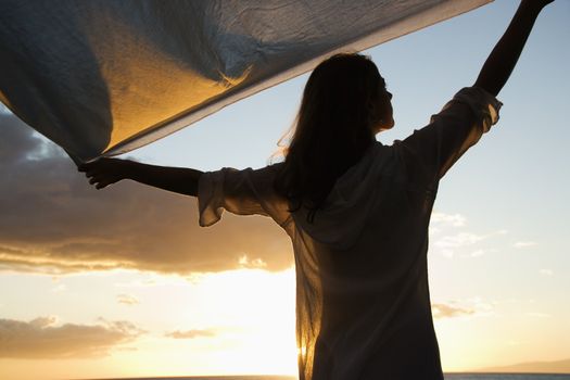 Attractive Caucasian mid-adult woman holding up fabric in breeze silhouetted by sunset beside ocean.