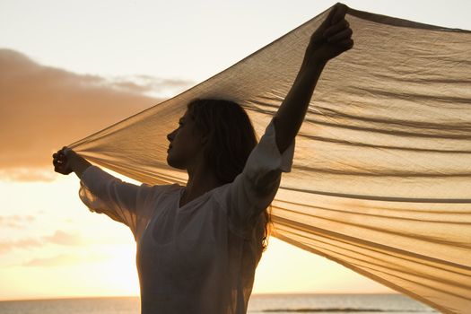 Attractive Caucasian mid-adult woman holding up fabric in breeze silhouetted by sunset beside ocean.