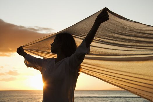 Attractive Caucasian mid-adult woman holding up fabric in breeze silhouetted by sunset beside ocean.