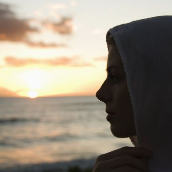 Pretty Caucasian mid-adult woman with scarf over head profiled against sunset over ocean.