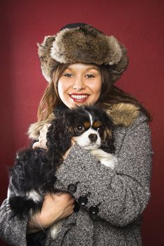 Young adult Caucasian woman wearing fur hat holding King Charles Spaniel in arms and smiling at viewer.