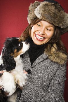 Young adult Caucasian woman wearing fur hat holding King Charles Spaniel in arms and smiling.