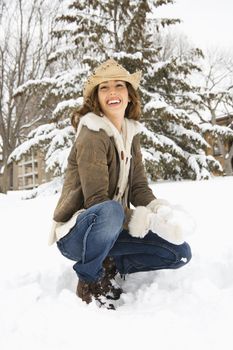 Caucasian young adult female smiling at viewer while kneeling in snow with snowball and wearing straw cowboy hat.