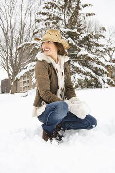 Caucasian young adult female looking over shoulder while kneeling in snow with snowball and wearing straw cowboy hat.