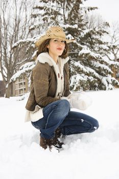 Caucasian young adult female looking at viewer while kneeling in snow with snowball and wearing straw cowboy hat.